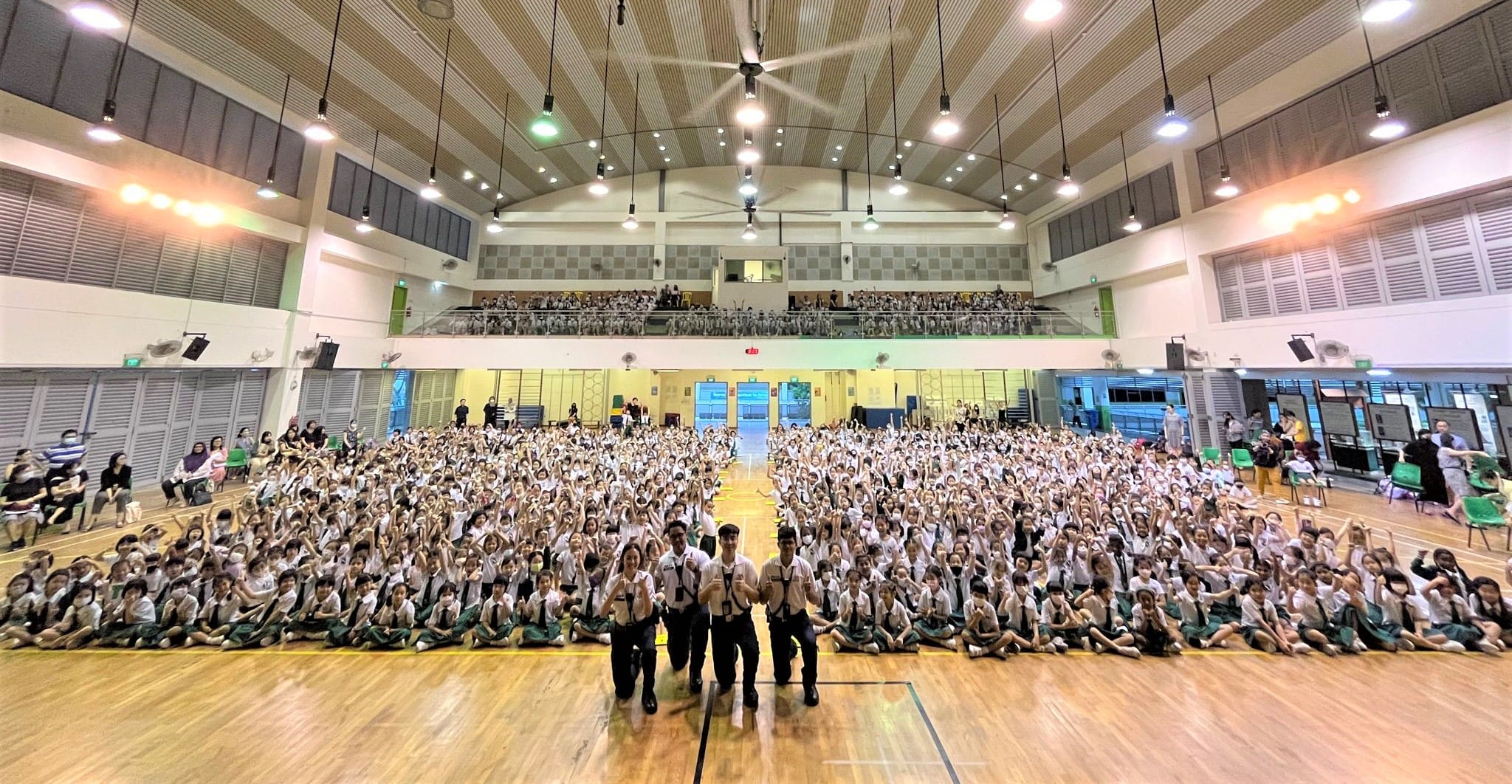 Large group of uniformed students sitting on a school gymnasium floor with adults standing at the front.